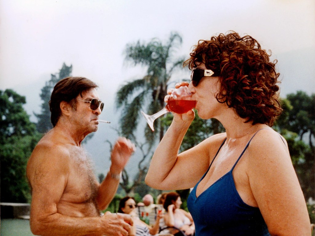 Fotograma de una mujer recostada en una silla de jardín, copa de vino en mano, rodeada de calor y desorden doméstico.