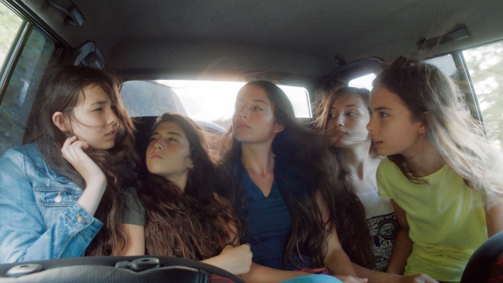 Las cinco hermanas protagonistas corren de la mano por un camino rural, riendo, con el cabello suelto y vestidos claros.