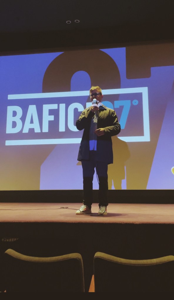 El director artístico del BAFICI, Javier Porta Fouz, hablando en el escenario de la Sala Lugones del Teatro San Martín durante el acto inaugural de la edición número veintisiete, con el micrófono y la pantalla de fondo.